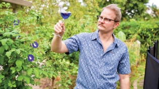 Erik Lowman next to his butterfly pea plant, holding a glass of Magic City Gin infused with butterfly pea and soursop juice. 