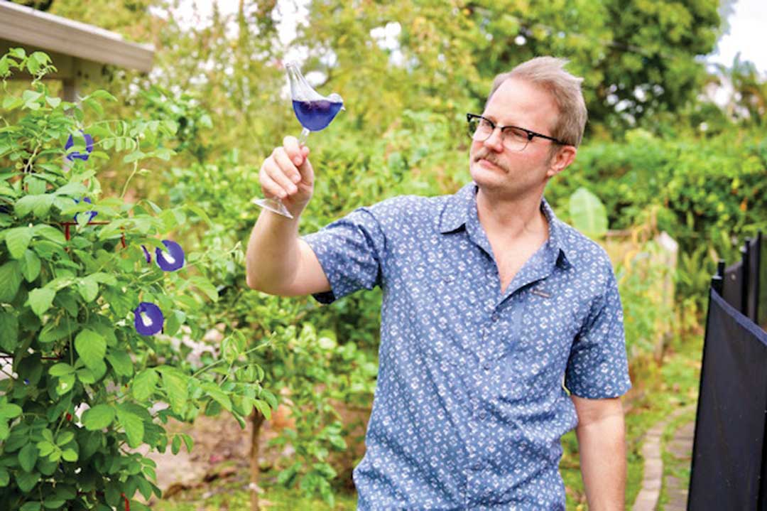 Erik Lowman next to his butterfly pea plant, holding a glass of Magic City Gin infused with butterfly pea and soursop juice.